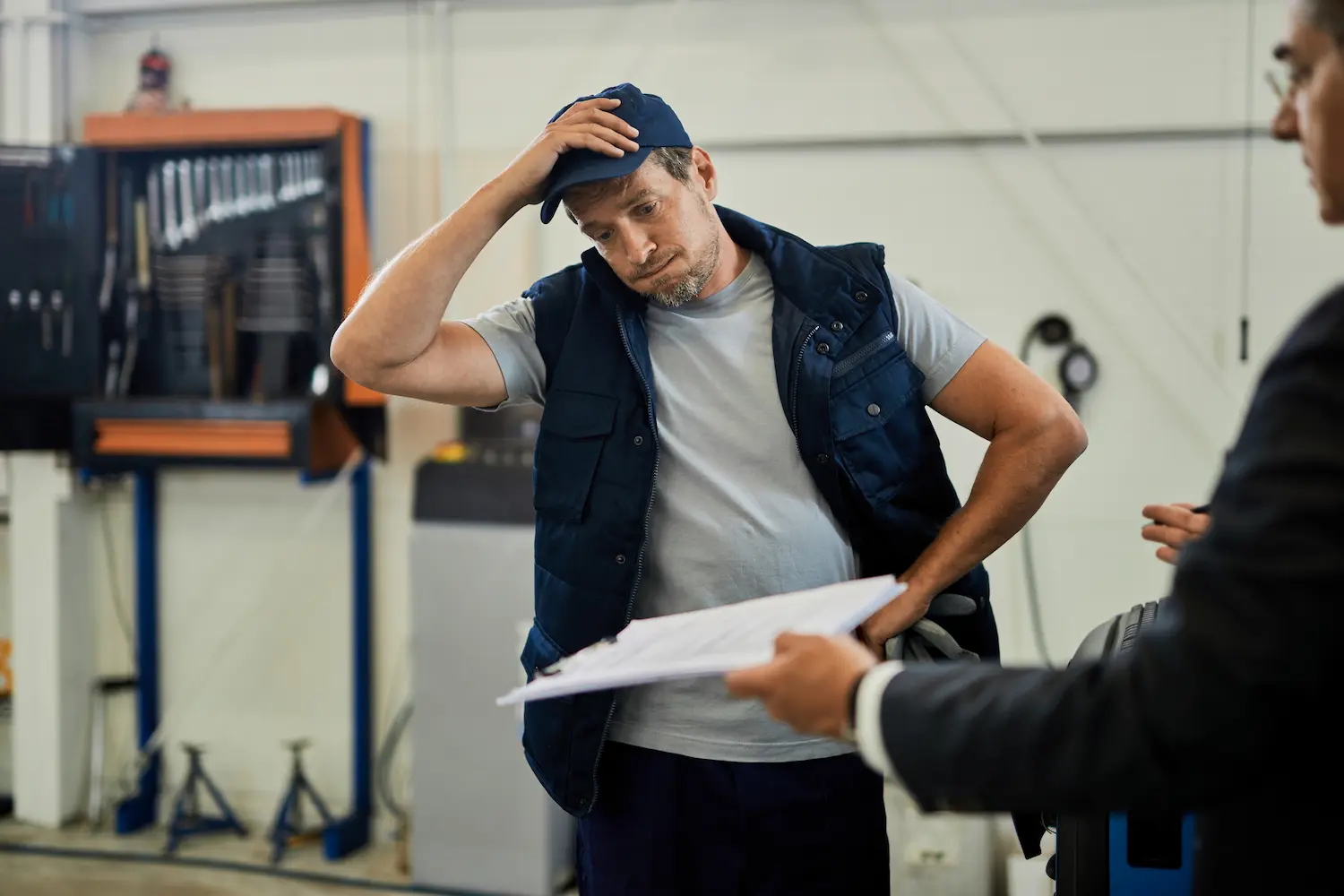 Mecánico estresado revisando papeles con un gerente vestido de traje dentro de un taller automotriz