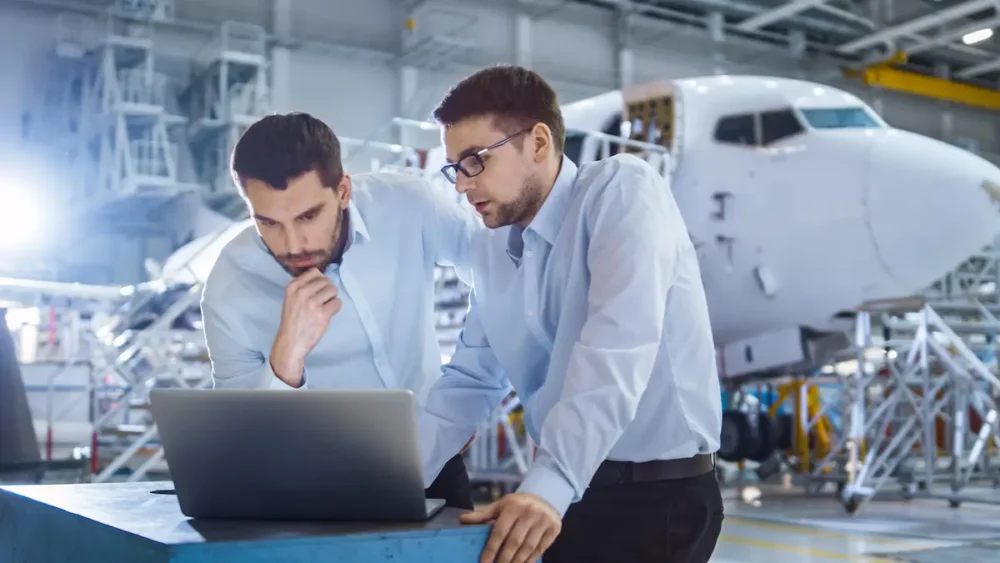 Dos ingenieros revisan un ordenador portátil en un hangar frente a un avión comercial en mantenimiento.