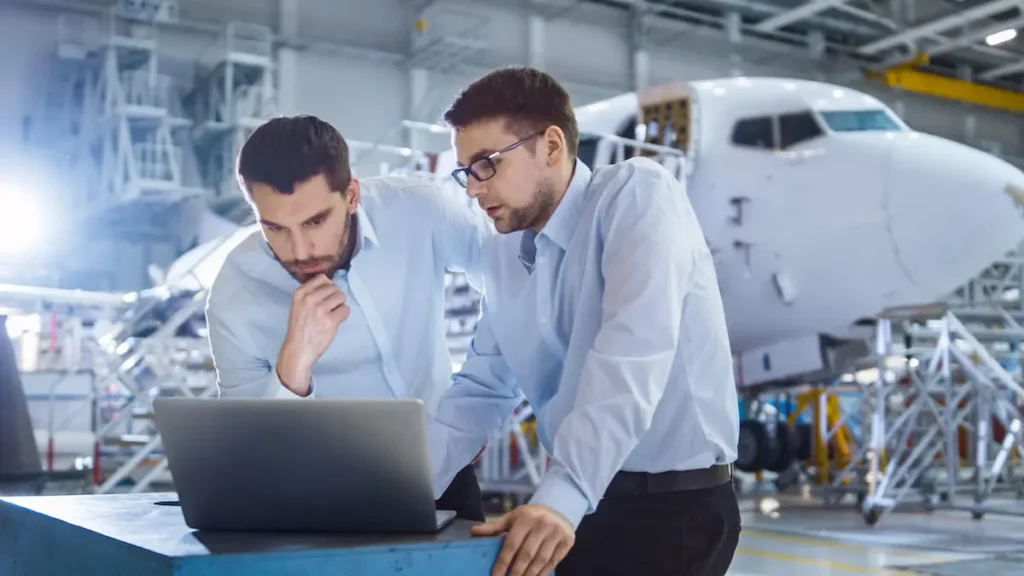 Dos ingenieros revisan un ordenador portátil en un hangar frente a un avión comercial en mantenimiento.