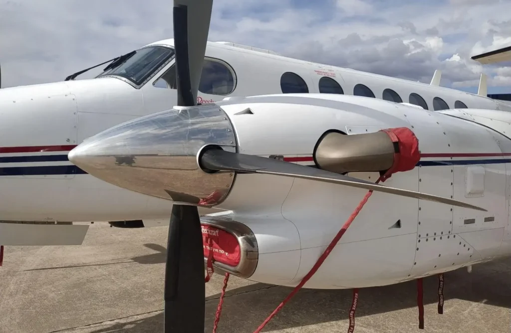 Motor y hélice metálica de avión Beechcraft blanco con cintas rojas de seguridad estacionado en pista.